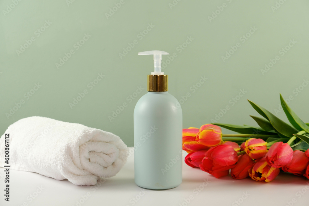 A green plastic bottle of cleaning agent with space for text and white moss twisted towel and red flowers on a white green background.