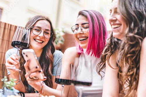 Group of happy friends drinking red wine enjoying happy hour sitting at winery bar- Three young girls having fun together watching smartphone outdoors at restaurant terrace-Youth culture concept 