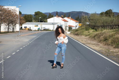 Young, pretty, brunette woman with curly hair, white top, jeans and heels, standing posing in the middle of a lonely road. Concept beauty, fashion, posing, model.