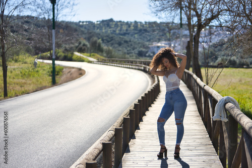 Young, beautiful, brunette woman with curly hair, white top, jeans and heels, sad and lonely, looking at the ground, standing in the middle of a wooden catwalk. Concept beauty, fashion, loneliness.