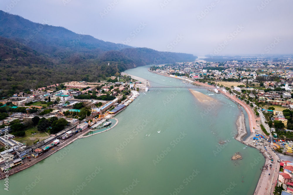 aerial drone shot of blue water of river ganga stretching into distance with himalayas with ram setu suspension bridge and temples on the banks of the river