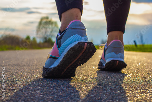Women's legs in running shoes on an asphalt road in the rays of the setting sun in summer. A woman goes in for sports by jogging in the countryside on an asphalt road. Running concept.