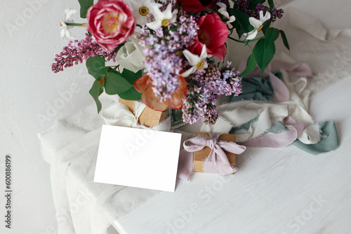 Greeting card mock up and lilac  tulips  daffodils flowers with present and pastel ribbons in modern rustic room. Happy Mothers day. Space for text. Stylish bouquet with gift box on wooden table