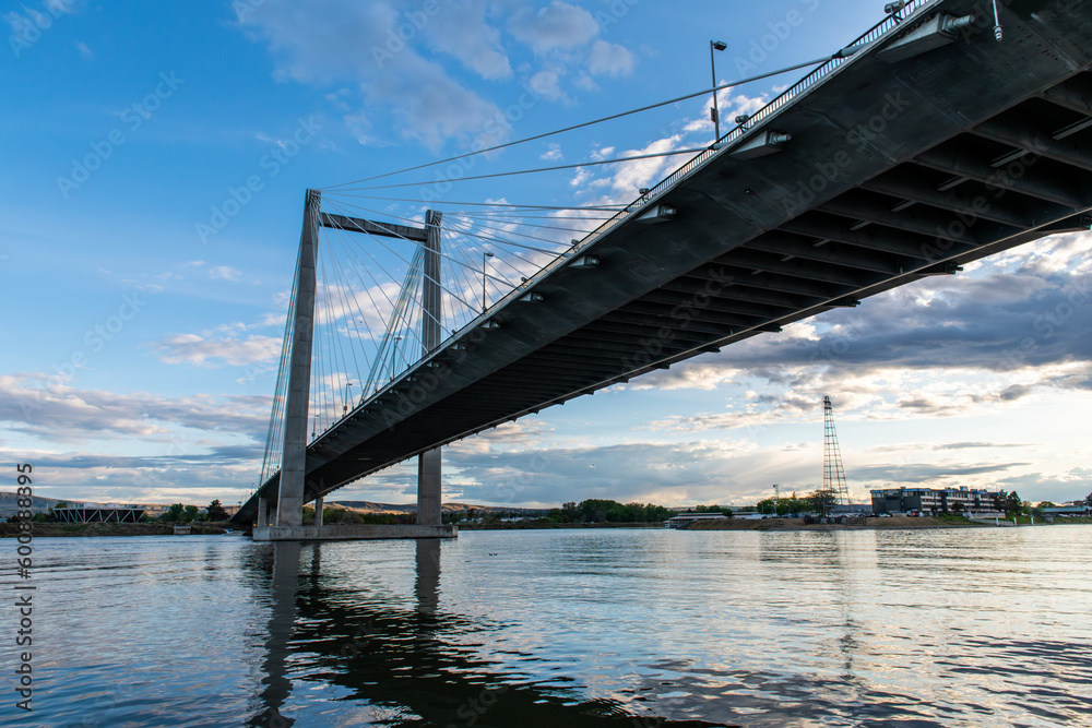 A cable bridge spans across a river against a cloudy blue sky at sunset.