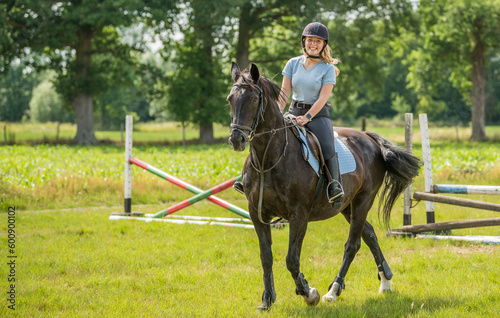 Young female adult girl riding hore on a meadow jumping obstacles and trees in the background.