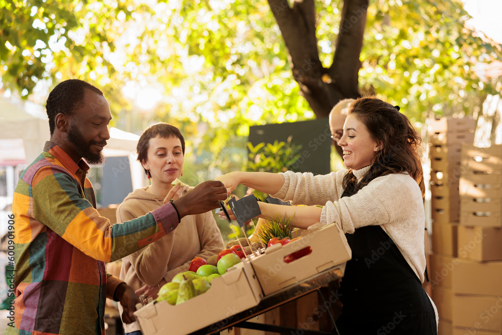 Vendor offering samples to customers while selling home-grown fruits and vegetables at local farmers market. Young multiracial family couple tasting natural organic produce while visiting food fair - obrazy, fototapety, plakaty 