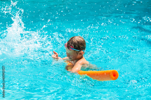 Oudoor summer activity. Concept of fun, health, vacation. Happy smiling boy five years old in swimming goggles laught and closes himself from splashing water in the pool with noodle on hot summer day.