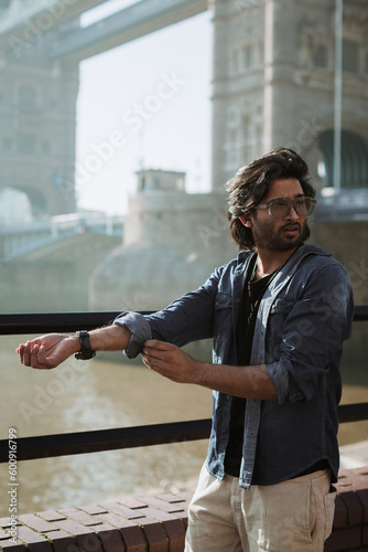 Close up handsome stylish guy in London is opposite of London Bridge, man standing on Tower Bridge. He wears a denim jacket, sunglasses and he is looking away from camera. photo