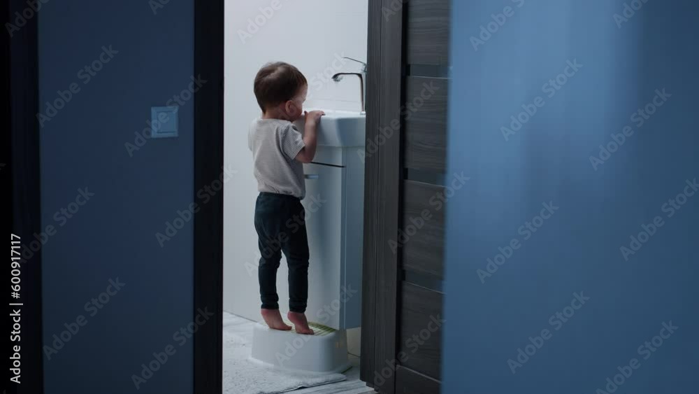 Baby boy stands on the stool to reach the sink. Curious kid observes how the water flows. Black cat enters the bathroom.