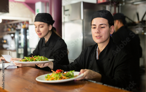 Focused young woman chef giving out finished dish at ordering station in open restaurant kitchen