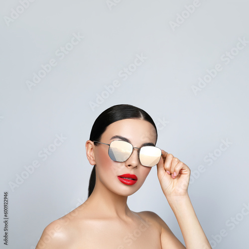 Attractive woman in stylish sunglasses on light grey background. Sky with clouds reflecting in lenses