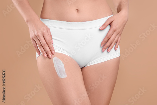 Woman with smear of body cream on her leg against light brown background, closeup
