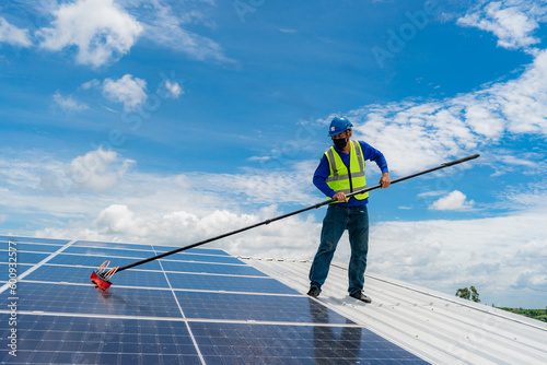 Professional worker cleaning solar panels with brush and washing with water on roof structure of building factory. Technician using mop to clean the dirty and dust, green electricity energy technology