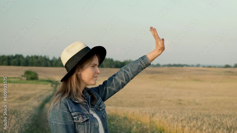 Girl is saying hello to someone by waving her hand up in the air. Woman is dressed with denim jacket and summer style hat. She looks like happy traveler on her holidays traveling around the country.