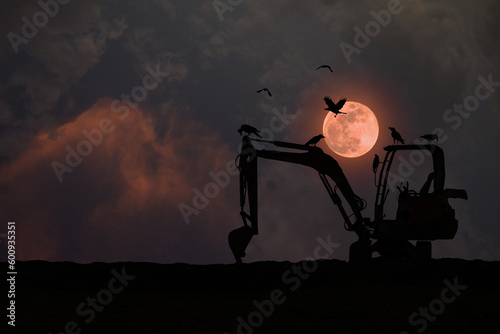 Silhouette of an excavator parked on the ground with a flock of crows at night with a full red moon.