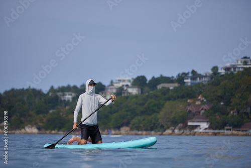 Guy in white hoodie standing on board on his knees, holding paddle in hands, floating in the calm water of tropical bay, searching for wave, happy holiday concept, copy space