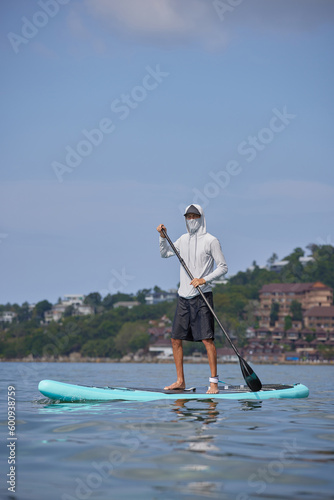 Man in white hoodie and mask standing with paddle in hands, floating on surf board with green coast on background, aquatic sport concept, copy space