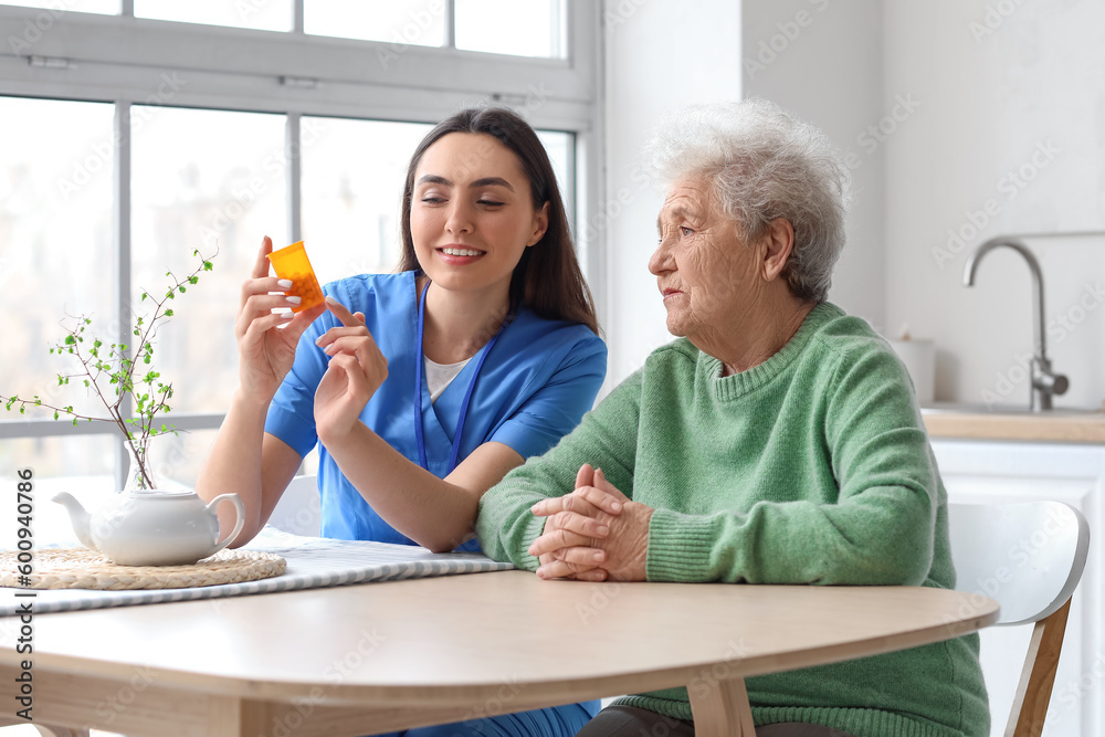 Young caregiver and senior woman taking pills in kitchen - obrazy, fototapety, plakaty 