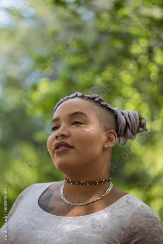 With a gentle smile, an African American and Latinx model stands in a forest. They express themes of optimism and connecting with nature, with a background of bright sunlit leaves.