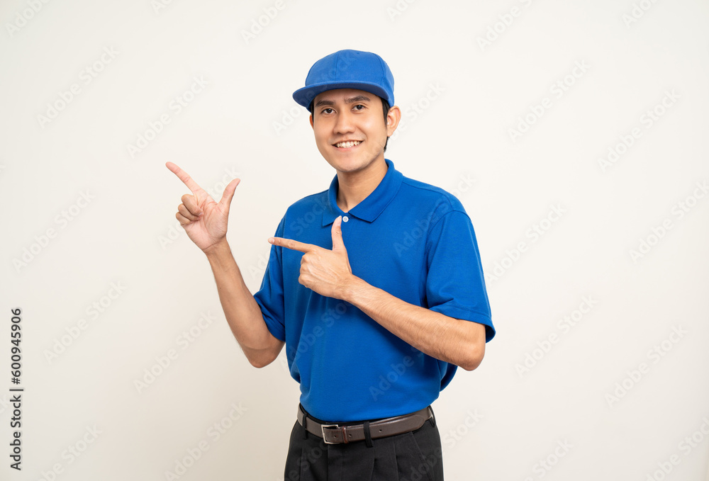 Happy delivery asian man in blue uniform standing on isolated white background. Smiling male service worker pointing the finger to blank space. Delivery courier and shipping service.