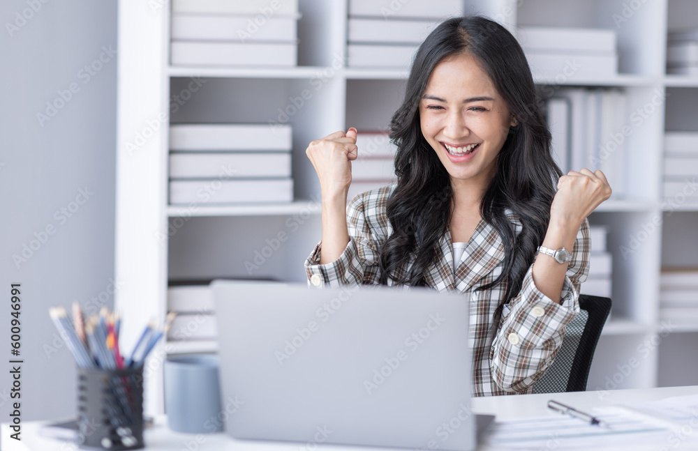 Excited Asian young woman using phone and laptop sitting on a desk office in the day at office Excited Asian young woman using phone and laptop sitting on a desk office in the day at office