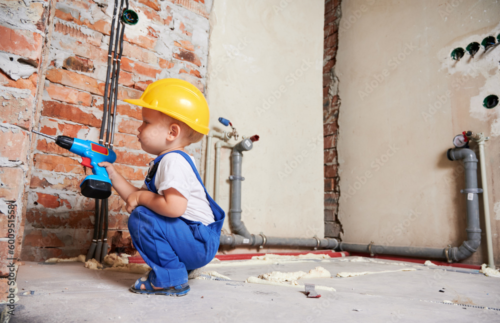 Kid construction worker crouching down and drilling brick wall with cordless electric drill. Baby boy in safety helmet using drilling power tool while working on home renovation.