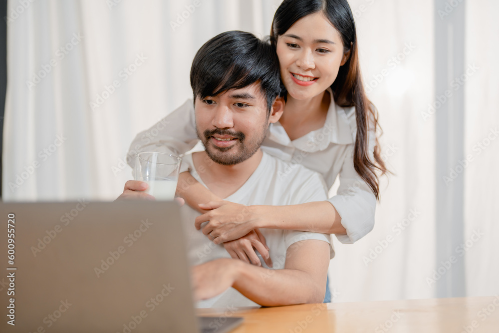 Happy young loving family couple looking at laptop computer screen, reading ecipes. Wife showing embrace to husband to pretty joyful husband, sitting at table in kitchen at home in weekend. Happy young loving family couple looking at laptop computer screen, reading ecipes. Wife showing embrace to husband to pretty joyful husband, sitting at table in kitchen at home in weekend.