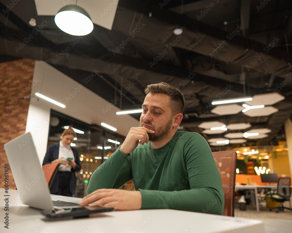 Bearded man holding his head while working on a laptop in an open space office. Red-haired caucasian woman uses smartphone. 