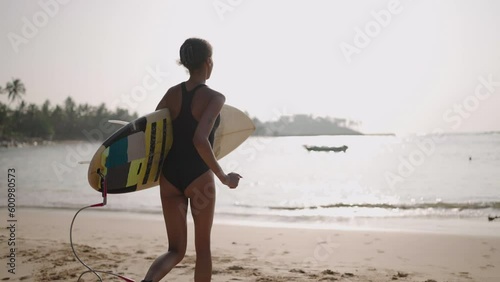 African woman running with surfboard in ocean water. Black female surfer with surf board. Pretty multiethnic girl goes on surfing session on tropical location spot at sunny sunrise.