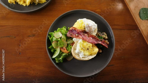 Breakfast is ready, woman put plate on table, split buns with poached eggs served with simple salad, pan-fried slice of bacon and mushrooms. Popular and tasty meal for morning time