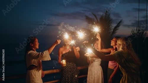 Diverse women dance, laugh with sparklers at seaside tropical hen party at sunset. Young smiling multiracial females have fun at 4th July Independence Day celebration by the sea with sparkling lights