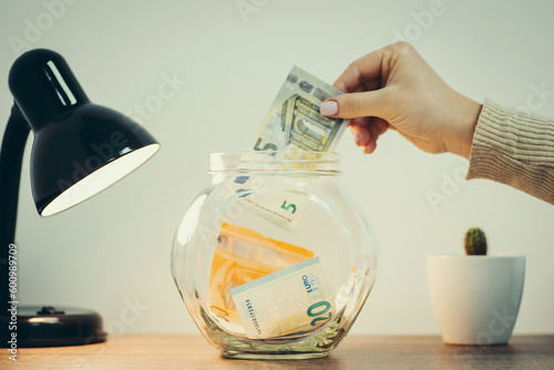 Female hand putting 5 Euro banknote to the glass jar. Saving money to buy something concept, toned image