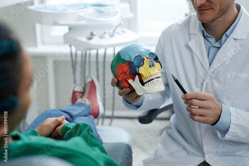 Closeup of professional male dentist holding skull model while consulting patient on tooth implantation technique, copy space