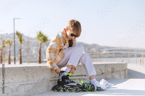 Caucasian blond preteen boy putting on roller skates sitting on the seafront. Hobby for kids, active lesure for teenagers. photo