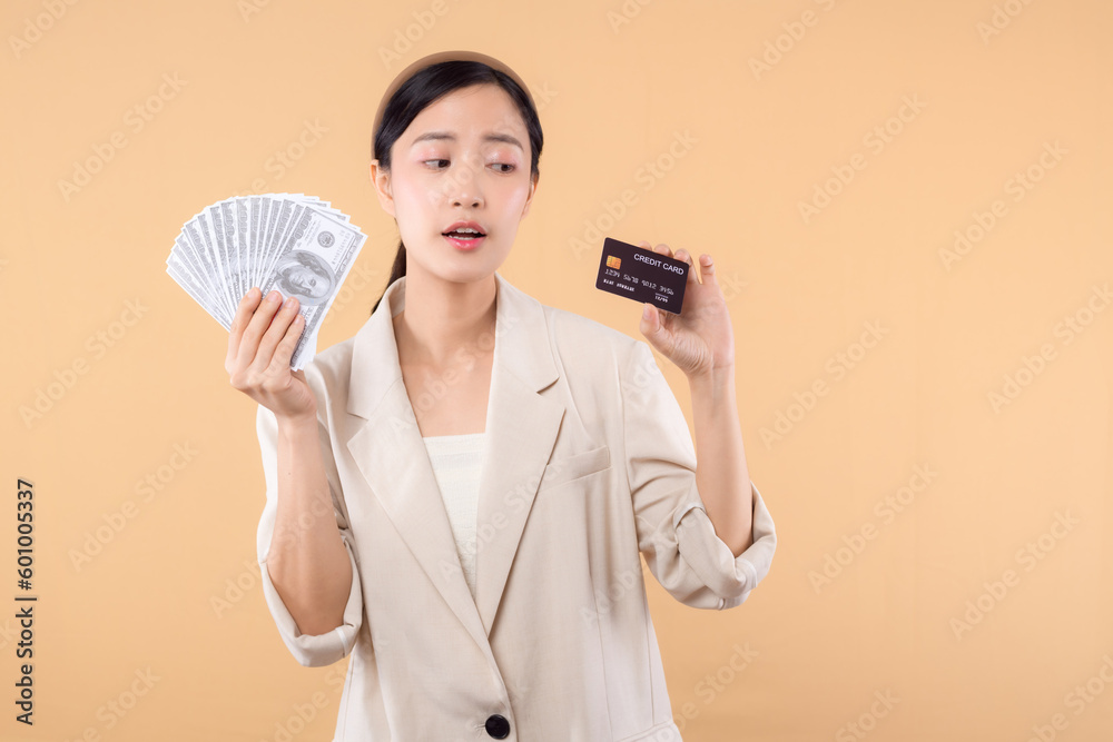 portrait of happy successful confident young asian business woman wearing white jacket holding cash money dollars and credit card standing over beige background. millionaire business, shopping concept portrait of happy successful confident young asian business woman wearing white jacket holding cash money dollars and credit card standing over beige background. millionaire business, shopping concept