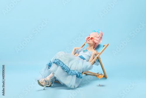 Portrait of pretty princess, queen wearing elegant dress sitting on sunbed and sunbathing over blue background. Summer time photo