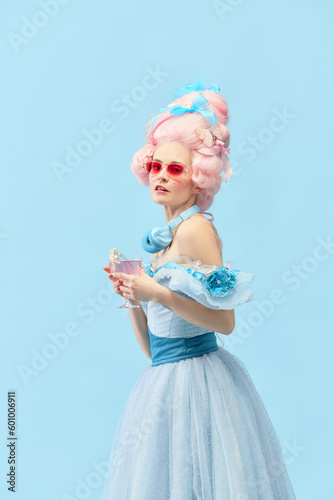 Portrait of young princess, queen wearing elegant dress, and pink wig drinking cocktail over blue studio background. Royal family party photo