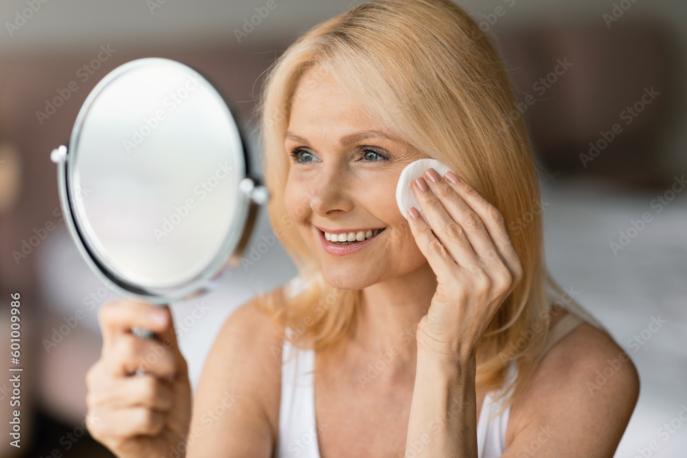 Aged woman cleansing skin with cotton pad, looking at mirror and smiling to her reflection, enjoying self care routine Aged woman cleansing skin with cotton pad, looking at mirror and smiling to her reflection, enjoying self care routine
