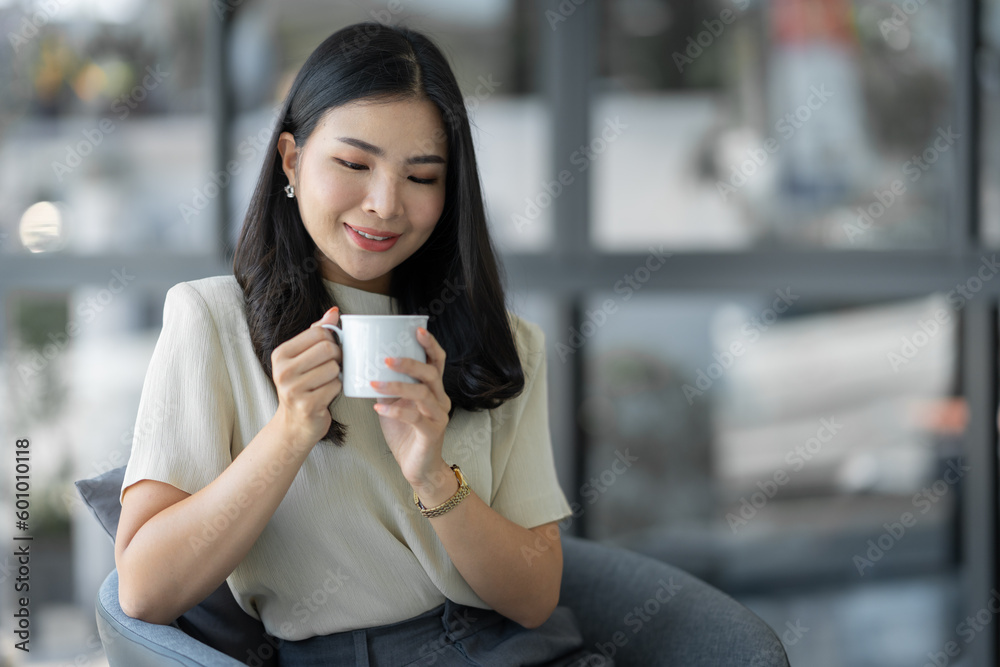 Attractive young Asian woman or businesswoman sitting drinking and holding cup of coffee relaxing, taking break while sitting cross legged on sofa chair at office. Attractive young Asian woman or businesswoman sitting drinking and holding cup of coffee relaxing, taking break while sitting cross legged on sofa chair at office.