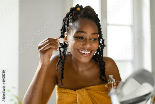 Luxurious self-care moments. Beautiful black lady applying skin serum on face with dropper  looking at mirror in bedroom
