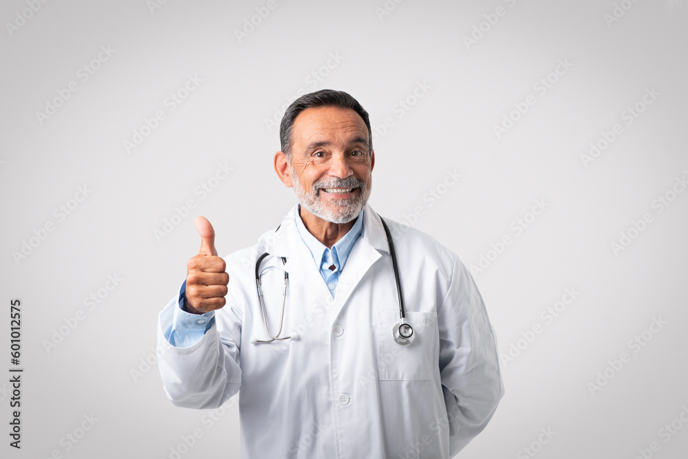 Smiling european senior man doctor in white coat show thumb up on gray background Smiling european senior man doctor in white coat show thumb up on gray background