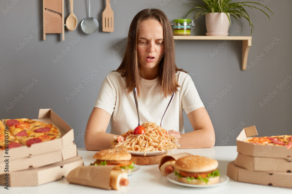 Confused shocked woman wearing white t- shirt siting at table with fast food in kitchen looking at unhealthy dishes with puzzled face, ordering too much meal.