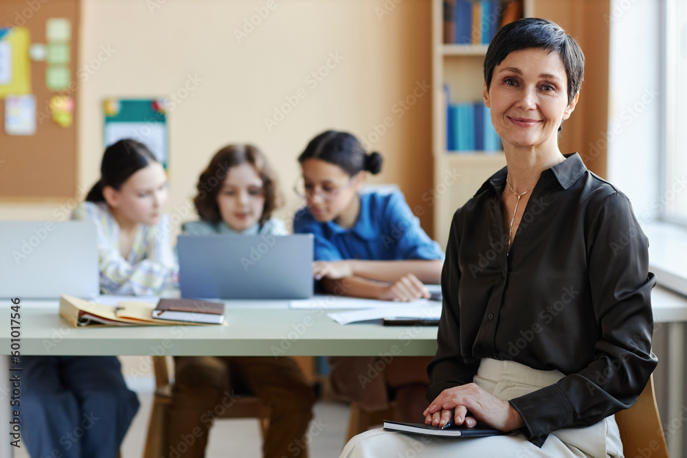 Portrait of mature teacher looking at camera sitting at lesson with children in background Portrait of mature teacher looking at camera sitting at lesson with children in background