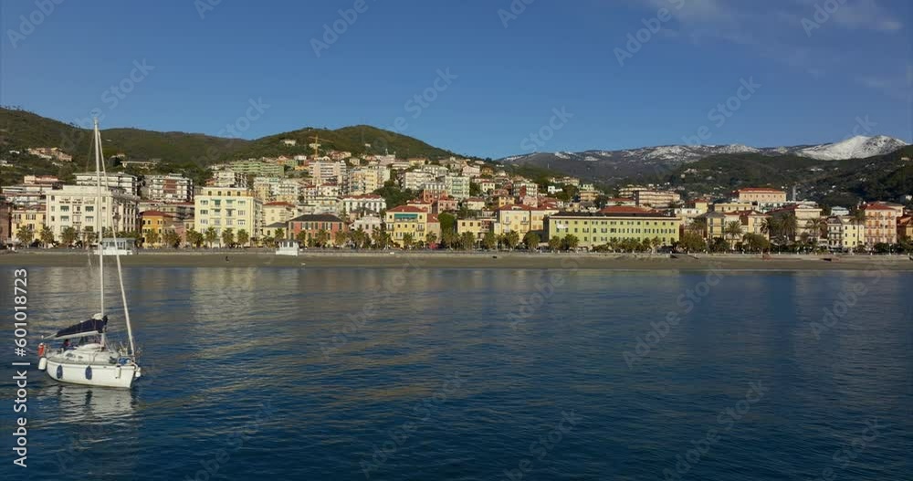 Aerial side view of white yacht sailing through calm ocean waters of Varazze in front of seaside village community and mountains in the background. Aerial side view of white yacht sailing through calm ocean waters of Varazze in front of seaside village community and mountains in the background.