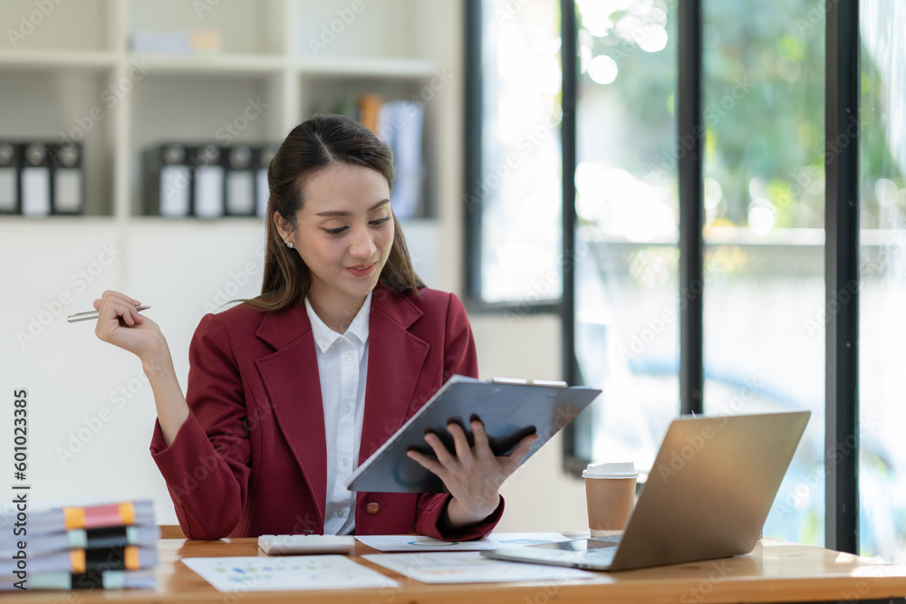© crizzystudio - Beautiful attractive Asian businesswoman holding a clipboard of documents sitting analytically analyzing various marketing management information the finances were at her desk in the office at ease.