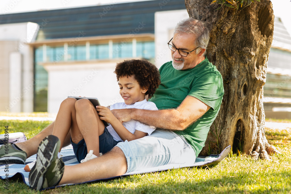 Smiling old caucasian man hug little mixed race boy, sit on green grass, enjoy free time, watch video Smiling old caucasian man hug little mixed race boy, sit on green grass, enjoy free time, watch video