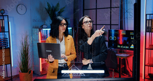 Pretty confident industrious high-skilled two businesswomen working together near glass wall in evening office,they recording important marks from computer report