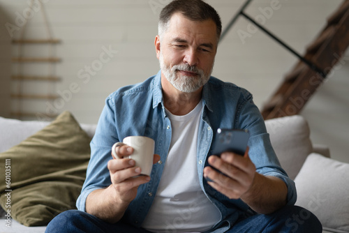Happy smiling senior man using smartphone device while sitting on sofa at home