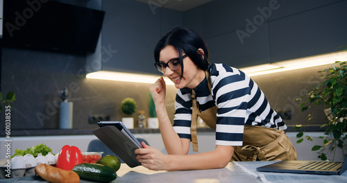 Attractive surprised dark-haired young woman showing emotional reaction on good news on tablet screen when standing in beautiful cuisine