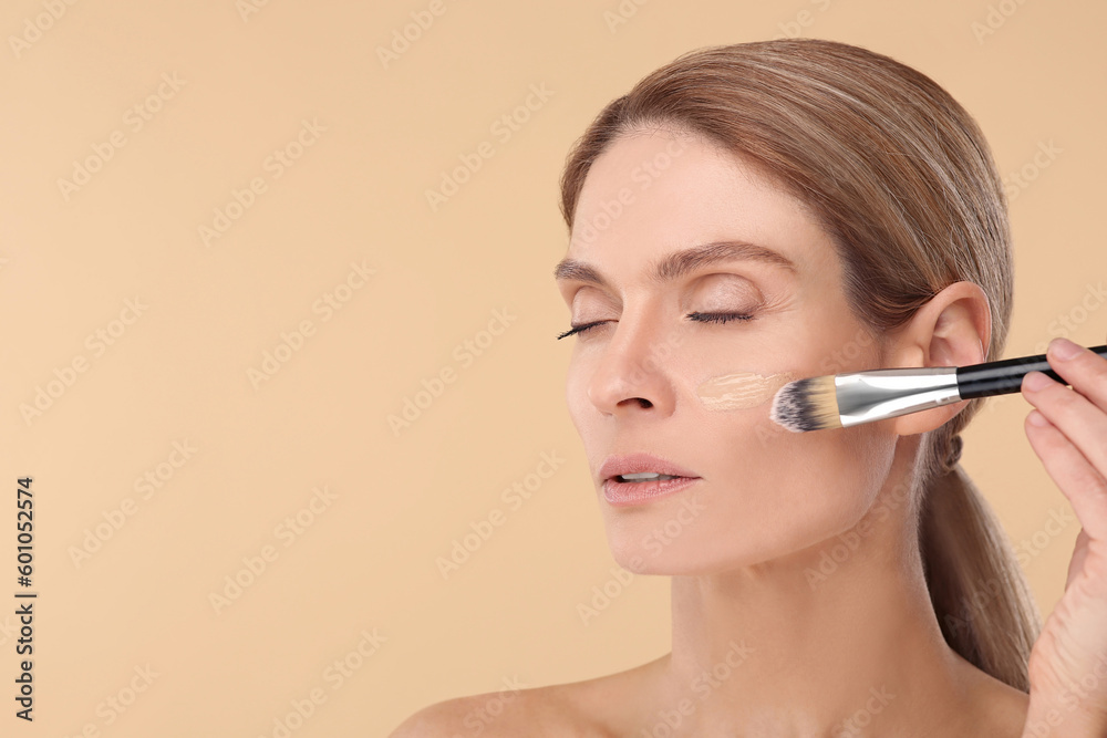 Woman applying foundation on face with brush against beige background. Space for text Woman applying foundation on face with brush against beige background. Space for text